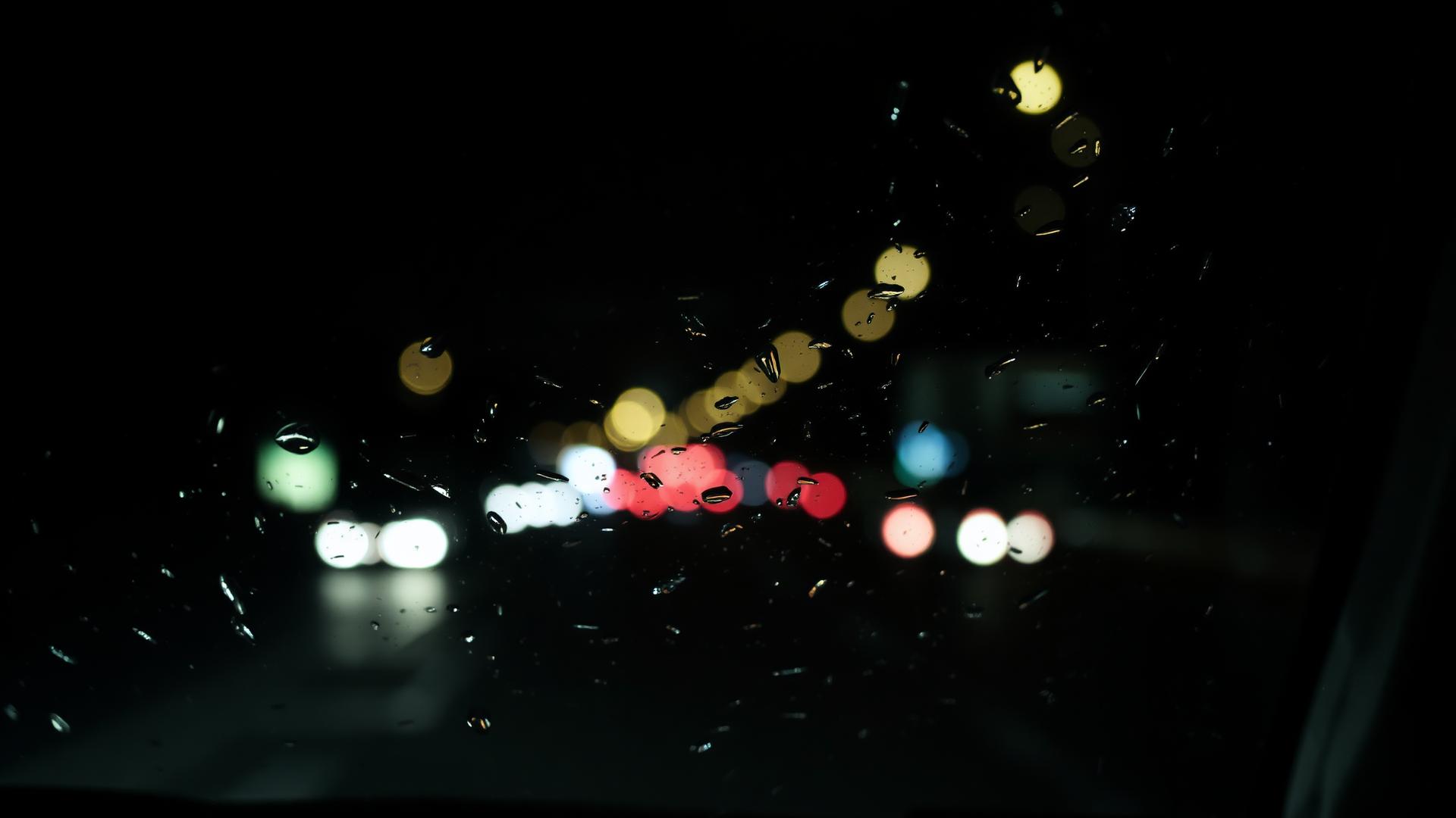 Pluie sur une vitre de voiture la nuit, lumières de ville floues à l’arrière-plan